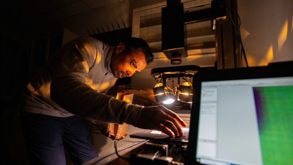 Man adjusting laboratory instruments in a dark room crouching behind a computer screen