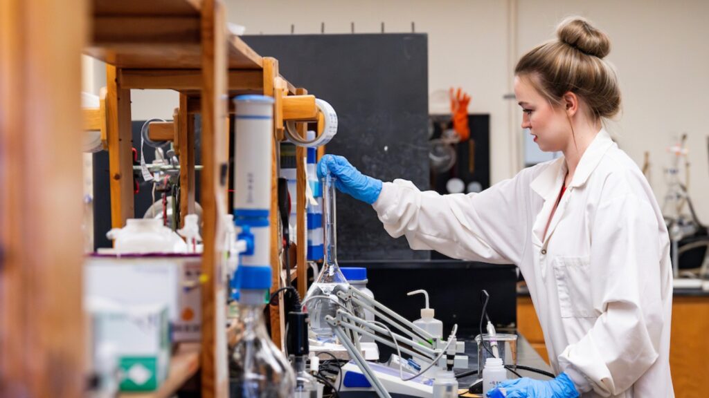 A woman in a lab coat with her hair in a bun reaching for glass equipment on a long wood shelf in a laboratory